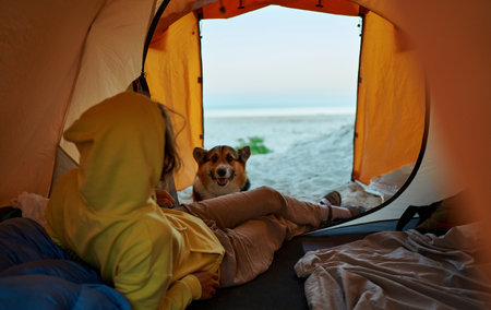 Female tourist relaxing in her tent, admiring breathtaking natural scenery. Charming Corgi dog gazes into the tent. Companionship and adventure to journeyの写真素材