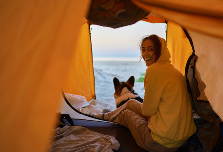 Happy young millennial girl with her puppy enjoying camping weekend and sitting inside orange tent. Freedom of traveling with beloved petsの写真素材
