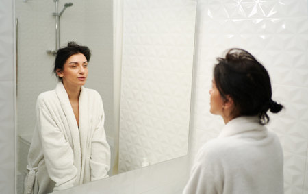 Tired woman in a robe standing in front of mirror, looking sleepy and exhausted. Concept of fatigue syndrome, sleep deprivation and morning routine.の写真素材