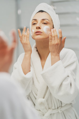 Woman using moisturizing foam for face washing, morning skincare routine. Adult female wearing towel on head looking in mirror in bathroom. Preparation for make upの写真素材