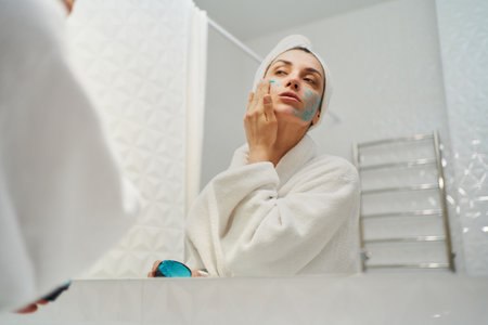 Rejuvenating power of natural skincare. woman wearing a robe while applying a nutritious healthy facial mask and looking in mirror in bathroomの写真素材