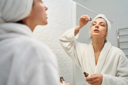 Stunning woman in bathrobe applying serum to her face as part of her daily skincare routine. Perfect for beauty and skincare related projectsの写真素材