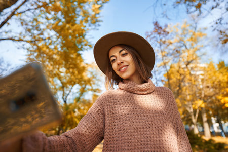 Woman in a chic hat and cozy knit sweater capturing perfect selfie moment on smartphone while strolling through sunny autumn parkの写真素材