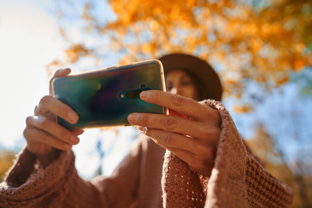 Close up smartphone in hands woman with sunlights on backdrop. Girl making photo on camera phone during autumn sunny dayの写真素材