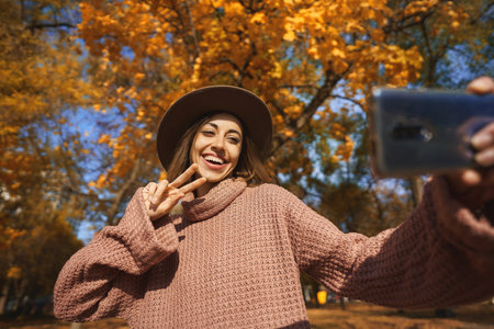 Playful smiling woman in treny hat and warm sweater posing for camera phone at fall season with orange foliage parkの写真素材
