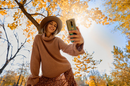 Fall fashion stunning image of woman in hat and cozy knit sweater taking picture on her smartphone at fall park with orange foliageの写真素材