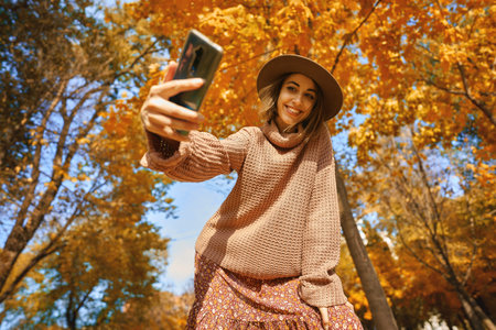 Autumn style with this trendy shot. Stylish woman in hat and knit sweater captures the moment with selfie on smartphone at park with orange leavesの写真素材
