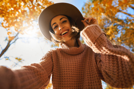 Cheerful smiling woman in hat making selfie at autumn sunny park. Bottom view of young lady happy positive emotionsの写真素材