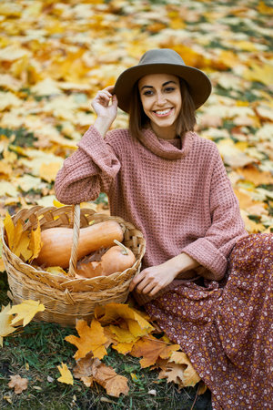 Fashionable woman in cozy knitted sweater, elegant dress, and chic hat sits beside a basket with pumpkins. Surrounded by golden autumn leaves on the groundの写真素材