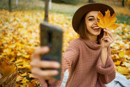 Smiling autumn woman in hat and knit sweater captures the moment with selfie on smartphone at park with maple leafe in handの写真素材