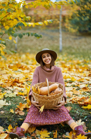 Young happy country woman in autumnal stylish wear holding wicker basket filled with freshly ripe pumpkins. Nature beauty of fall season and autumnal harvestの写真素材