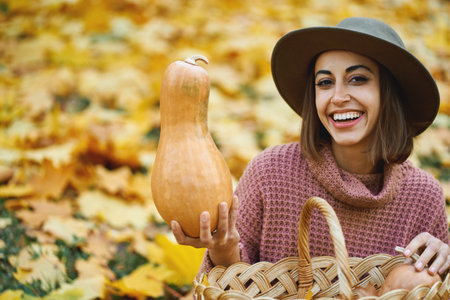 Close up autumn portrait of happy smiling lady in hat holding big rape pumpkins outdoors, fall season and colorful foliage around on backdropの写真素材