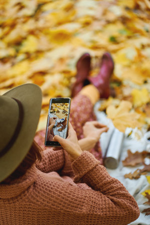 Rear view of fashionable woman in hat taking photo on camera phone while relaxing in beautiful autumn park. Focus on picture in smartphoneの写真素材