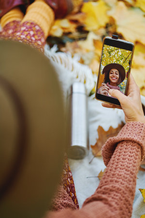 Top view of woman in hat taking selfie on her phone while enjoying time in cozy autumn park. Focus on picture in smartphoneの写真素材