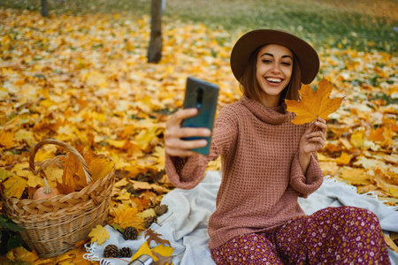 Autumn trendy style woman in hat and knit sweater captures the moment with selfie on smartphone at park with maple leafe in handの写真素材