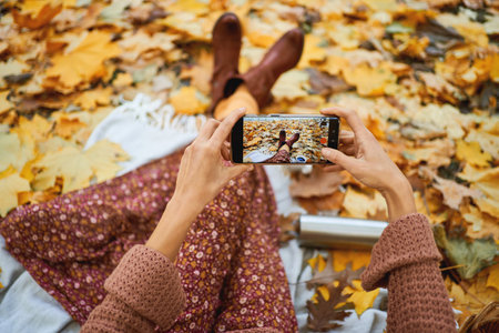 Top view of stylish autumnal woman taking photo of her legs on smartphone during picnic surrounded of orange leavesの写真素材