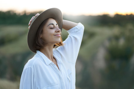 Graceful young lady in hat enjoying the beauty of nature amidst serene hillside landscapes during a captivating sunsetの写真素材