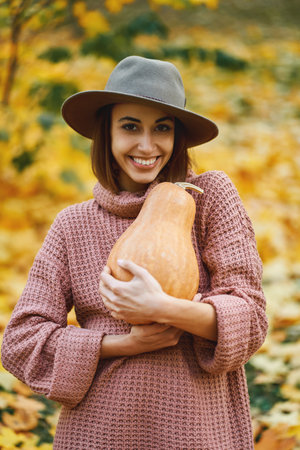 Vertical portrait pretty woman with charming smile wearing autumnal stylish clothes holding big pumpkin surrounded orange leavesの写真素材