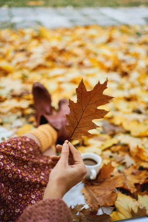 Trendy autumn fashion and atmospheric mood. Women in red dress and vintage boots lying surrounded fall leaves, holding leaf in handの写真素材