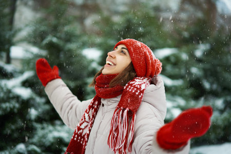 Young happy girl in red beanie and scarf standing in forest at snowy winter day outdoors. joyful looks up at snowflakes.の写真素材