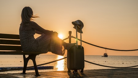 Romantic woman sitting by the sea with a travel bag at sunset. A ship is visible in the distanceの写真素材