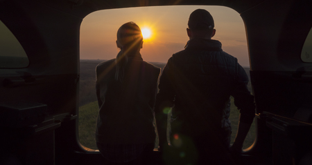A young couple sits next to the trunk of a car, admiring the beautiful sunset in a picturesque place in the mountainsの写真素材