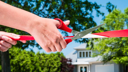 Hand with scissors cuts red ribbon in the background of new house