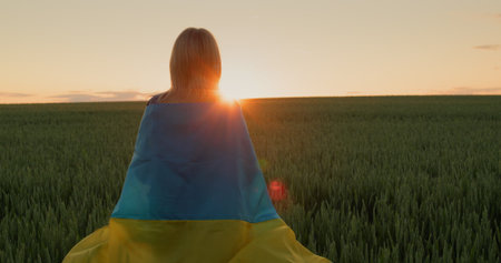 A woman with the flag of Ukraine on her shoulders looks at the sunrise over a field of wheat. Stand with Ukraine conceptの写真素材
