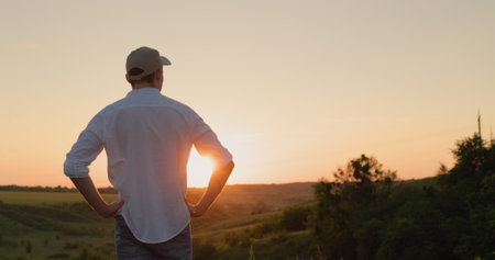 A confident young farmer stands in front of a picturesque countryside as the sun sets. View from the backの写真素材