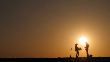 Against the backdrop of the setting sun, a woman and a man, one with a box of seedlings and the other with a tablet, talk about the sowing season as farmers in the field.の写真素材
