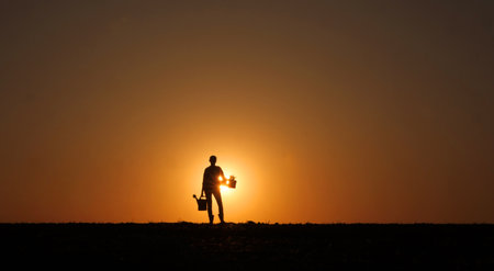 At sunset, a woman farmer stands in a field, her silhouette distinct as she holds a watering can and a seedling.の写真素材