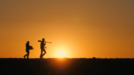 Two farmers with work equipment walk through a plowed field at sunsetの写真素材