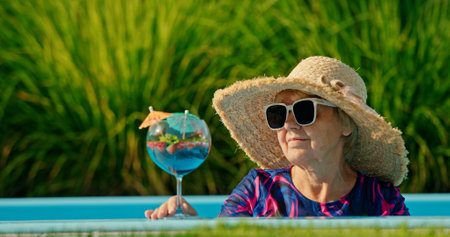 Elderly woman with a delighted expression, wearing a sun hat and sipping a cold drink while lounging in a pool on a bright dayの写真素材