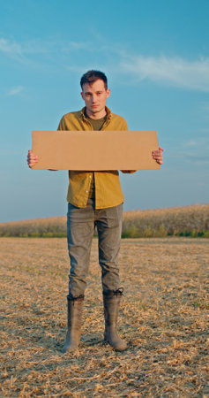 A man standing in a field holding a blank cardboard sign, wearing a yellow shirt and boots under a clear blue sky.の写真素材