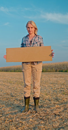 A woman stands in a field holding a blank cardboard sign, wearing a plaid shirt and boots under a clear blue sky.の写真素材