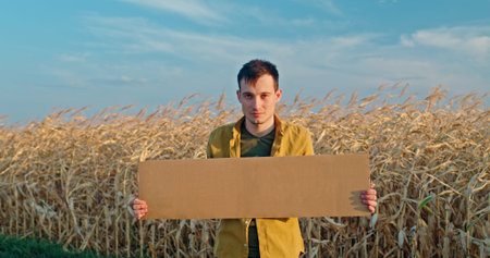 A man holding a blank cardboard sign stands in a dry cornfield, wearing a yellow shirt against a...