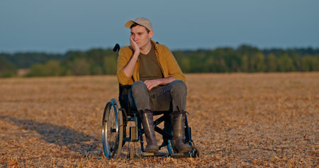 A man in a wheelchair sits in an empty field, resting his head on his hand, looking thoughtful.の写真素材