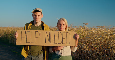 A man and a woman holding a HELP NEEDED sign in a field of dry crops, looking serious.の写真素材