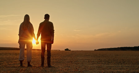 A young farming couple stands in a field, holding hands at sunset, while a combine harvests in the...
