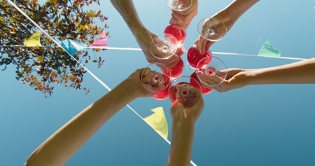 Six people toast with red drinks under a clear sky, surrounded by festive flags and sunlit leaves. Low angle viewの写真素材