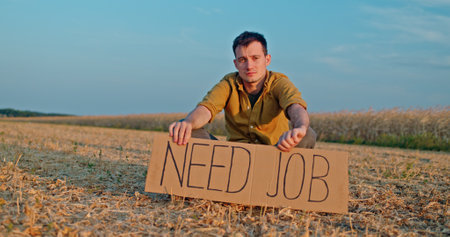 A young farmer sits in a field holding a sign that reads NEED JOB, reflecting employment challenges for farmers.の写真素材