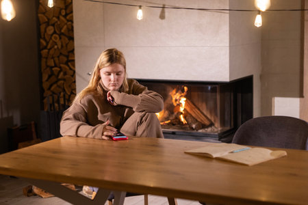 Teenage girl using a smartphone at a wooden table in a cozy room with a lit fireplace and warm string lights.の写真素材
