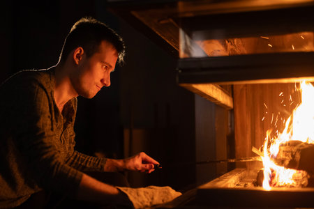 A young man carefully tends to a fireplace, adjusting the flames with a poker. The warm glow of the fire illuminates his face, creating a cozy and focused atmosphere.の写真素材