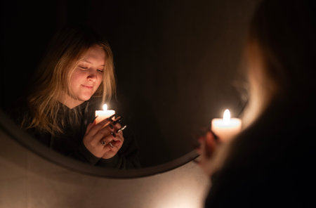 A young woman holds a lit candle, gazing softly at its flame. Her reflection is visible in a mirror,...