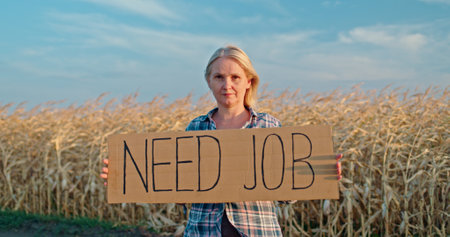 A woman stands before a golden cornfield, holding a Need Job sign. The image captures the essence of rural unemployment and determination, with a serene blue sky as the backdrop.の写真素材