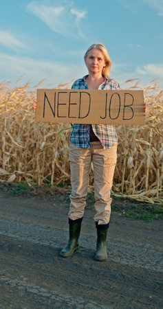 A middle-aged woman standing on a rural dirt road holding a cardboard sign reading NEED JOB. She wears outdoor work clothes, including a plaid shirt, khaki pants, and green bootsの写真素材