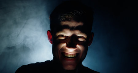 A man in the dark with a disturbing, wide grin, lit from below with dramatic shadows, evoking an eerie and haunting atmosphere. A concept of fear and suspense.の写真素材