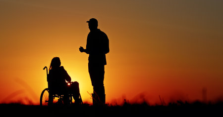 Silhouette of a standing person and a wheelchair user in a warm sunset. The scene conveys themes of companionship, support, and resilience in a serene rural setting.の写真素材