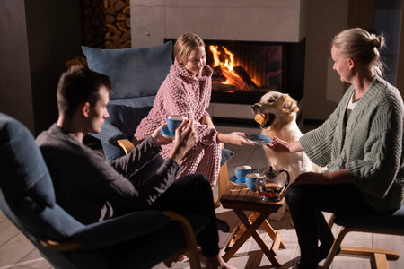 Family relaxing by a cozy fireplace, sharing tea and treats, with a Golden Retriever happily eating a snack in a warm, homely atmosphere.の写真素材