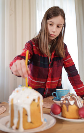 A young girl carefully placing a candle into an Easter cake covered with white icing and colorful decorations, with another decorated cake and a blue cup in the background.の写真素材